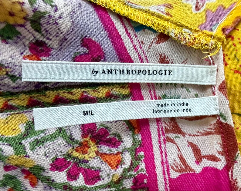 May include: A close-up of patterned fabric in pink, yellow, and purple with floral designs. Two white labels are visible, one with "by ANTHROPOLOGIE" and the other with "M/L" and "made in india / fabrique en inde".