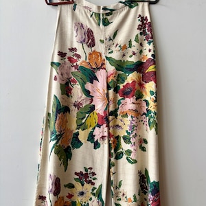 K&ouml;nnte beinhalten: Hoch taillierte Hose mit weitem Bein aus cremefarbenem Stoff mit einem lebendigen, mehrfarbigen Blumenmuster in Rosa-, Gelb-, Rot- und Gr&uuml;nt&ouml;nen. Die Hose hat einen geraden Schnitt und ist an zwei Metallklammern aufgeh&auml;ngt.