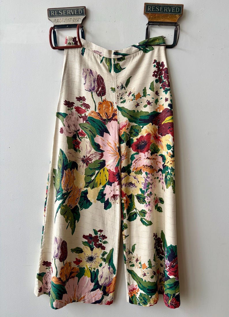 K&ouml;nnte beinhalten: Hoch taillierte Hose mit weitem Bein aus cremefarbenem Stoff mit einem lebendigen, mehrfarbigen Blumenmuster in Rosa-, Gelb-, Rot- und Gr&uuml;nt&ouml;nen. Die Hose hat einen geraden Schnitt und ist an zwei Metallklammern aufgeh&auml;ngt.