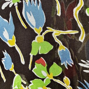 Peut inclure: Gros plan sur un tissu &agrave; motifs floraux. Le motif pr&eacute;sente des fleurs bleues, rouges et blanches avec des feuilles vertes et des tiges jaunes sur un fond marron fonc&eacute;. Les fleurs sont stylis&eacute;es.