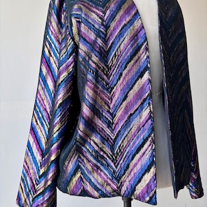 May include: A vintage open-front jacket with a striking chevron pattern. The jacket features a mix of colors, including blue, purple, gold, and black. The fabric has a metallic sheen, adding to its visual appeal. The jacket is displayed on a mannequin.