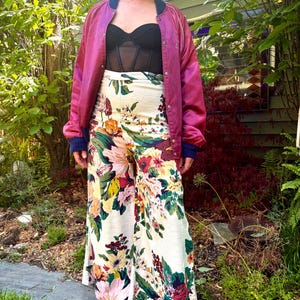 K&ouml;nnte beinhalten: Eine magentafarbene Satin-Bomberjacke mit dunkelblauem Rippkragen und B&uuml;ndchen, getragen &uuml;ber einem schwarzen tr&auml;gerlosen Korsett-Top und einer hoch taillierten weiten Hose mit buntem Blumenmuster auf cremefarbenem Grund.