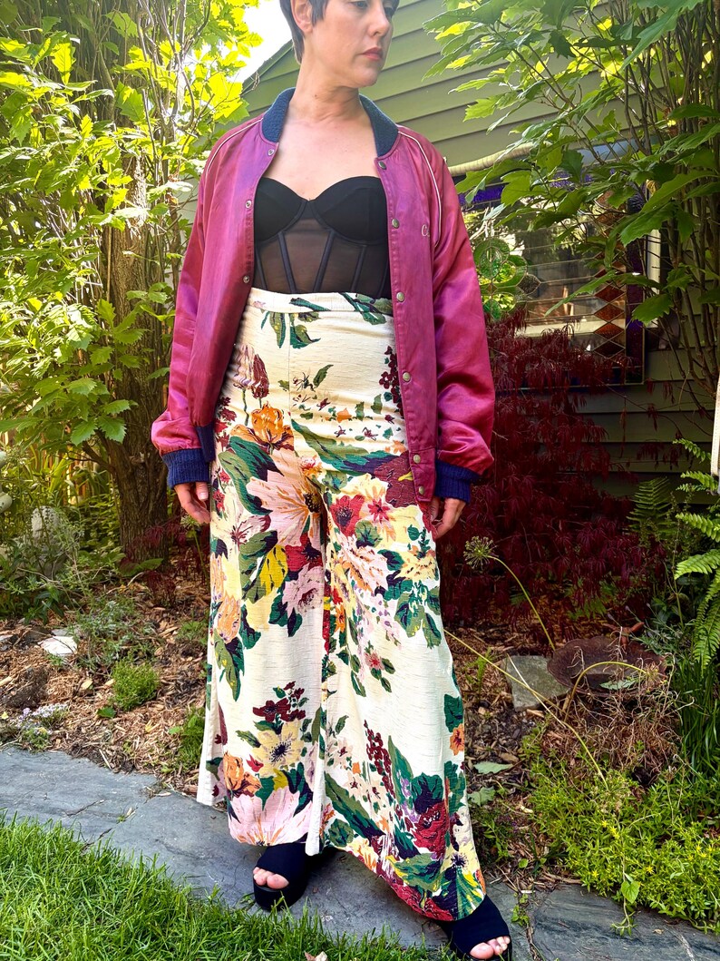 K&ouml;nnte beinhalten: Eine magentafarbene Satin-Bomberjacke mit dunkelblauem Rippkragen und B&uuml;ndchen, getragen &uuml;ber einem schwarzen tr&auml;gerlosen Korsett-Top und einer hoch taillierten weiten Hose mit buntem Blumenmuster auf cremefarbenem Grund.