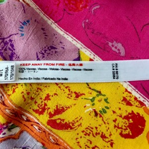 May include: A white tag with black text, including "KEEP AWAY FROM FIRE" and "100% Viscose", is placed over a colourful fabric with floral patterns in pink, yellow, and purple. The tag also includes text in multiple languages.