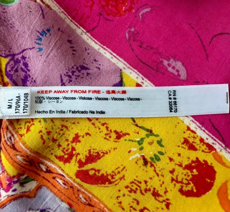 May include: A white tag with black text, including "KEEP AWAY FROM FIRE" and "100% Viscose", is placed over a colourful fabric with floral patterns in pink, yellow, and purple. The tag also includes text in multiple languages.