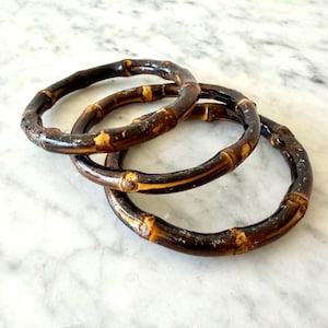 Peut inclure: Trois bracelets circulaires fabriqués à partir de segments de bambou brun foncé. Chaque bracelet présente la texture naturelle et noueuse du bambou avec des nœuds brun clair et une finition polie et brillante, disposés en couches.