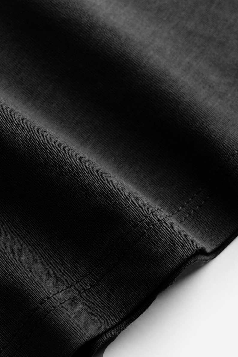 May include: Close-up of a black fabric, likely a garment, with visible texture and stitching along the edge. The fabric appears to be a knit material, possibly cotton or a blend, with a subtle sheen.