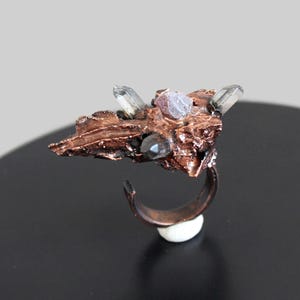 May include: A copper ring with a rough, organic design. The ring is adorned with clear quartz crystals and a black stone.