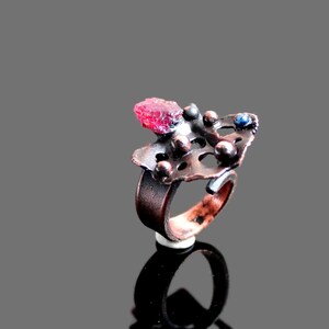 May include: A copper ring with a red gemstone and a blue gemstone set in a textured metal design.