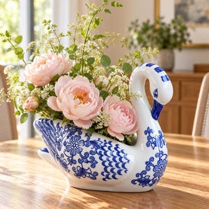May include: A white ceramic swan-shaped vase featuring intricate blue floral and scale patterns. The vase is filled with a bouquet of light pink peonies and delicate green filler flowers, creating a decorative centrepiece for a wooden table.