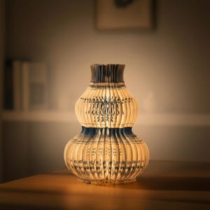 Op de afbeelding: Een decoratieve, dubbel kalebasvormige lamp met een blauw en wit bloemmotief. De lamp is gemaakt van papier of een vergelijkbaar materiaal, met een geplooide of gevouwen constructie. De lamp wordt van binnenuit verlicht en geeft een warme gloed. De lamp staat op een houten oppervlak.