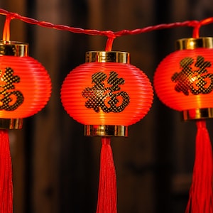 May include: A string of three bright red spherical lanterns connected by a red wire. Each lantern features a gold-colored top and bottom cap, a long red tassel hanging from the base, and a black Chinese character printed on the front surface.