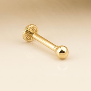 Pode incluir: Um piercing de nariz de ouro com uma pequena bola na ponta. O piercing é feito de ouro 14 quilates e tem 6 mm de comprimento.