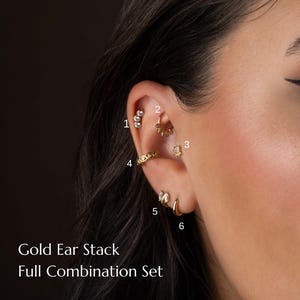 May include: A close-up of an ear adorned with six pieces of gold jewelry. The ear piercings include a triple stud, a hoop with charms, a small stud, a chain-like ear cuff, a small hoop with a gem, and a simple gold hoop. Text reads: Gold Ear Stack Full Combination Set.
