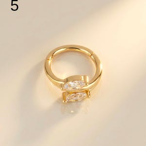 以下が含まれることがあります： 透明なマーキス型の宝石が付いた、金色のフープイヤリング。 イヤリングはヒンジ付きのデザインで、滑らかで光沢のある仕上がりです。 数字の「5」が左上にあります。