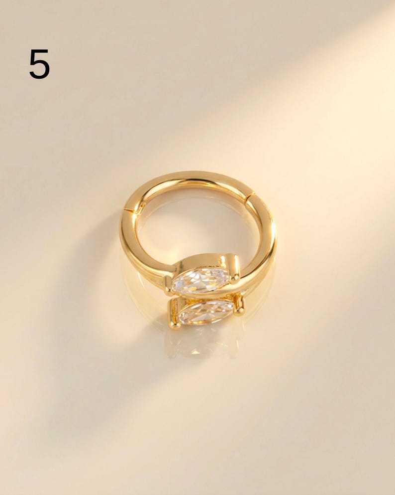 以下が含まれることがあります： 透明なマーキス型の宝石が付いた、金色のフープイヤリング。 イヤリングはヒンジ付きのデザインで、滑らかで光沢のある仕上がりです。 数字の「5」が左上にあります。