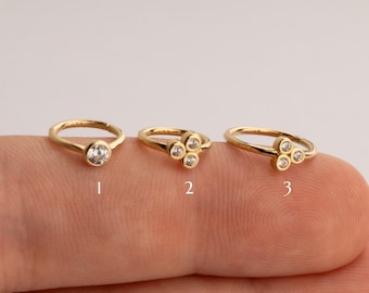 May include: Three gold nose rings with clear cubic zirconia stones. The first ring has one large stone, the second ring has four stones in a cluster, and the third ring has three stones in a cluster.