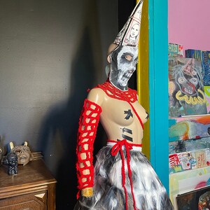 May include: A mannequin wearing a red crocheted top and skirt with a white and black painted design. The mannequin is wearing a white cone hat with the word "MAYBE" written on it. The mannequin is also wearing a white and black skull mask.