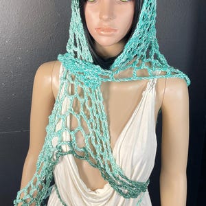 May include: A turquoise crocheted head covering and scarf set. The open-weave design is draped over a mannequin wearing a white dress. The scarf is tied at the waist with a matching turquoise cord.