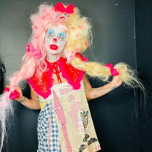 May include: A person with a pink and blonde wig styled with pink bows and a small doll head accessory. They wear a red lace collar over a patchwork dress featuring blue gingham, striped, and patterned fabric panels with printed bat graphics.