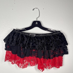 May include: A layered mini skirt featuring a black velvet top tier with a tonal floral pattern, a middle tier of black lace, and a bottom tier of vibrant red lace. The skirt is displayed on a black plastic hanger against a plain wall.