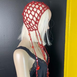 May include: A burgundy crochet head covering with a net-like design, adorned with red beads and dangling strands. The headpiece is displayed on a mannequin with blonde hair, showcasing its intricate details and texture.