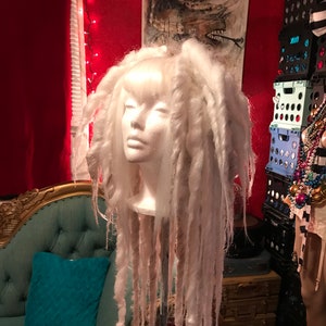Puede incluir: Una peluca blanca con rastas largas y gruesas. La peluca est&aacute; en una cabeza de poliestireno y est&aacute; sentada en un soporte.