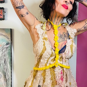 May include: A woman wearing a white crocheted dress with yellow crocheted straps and a crown. She has tattoos on her arms and a nose ring.