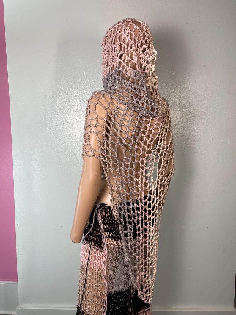 May include: A handmade crochet outfit in shades of pink, gray, and black. The outfit includes a hooded shawl and a skirt. The open weave design creates a net-like appearance. The mannequin is positioned against a gray and pink wall.