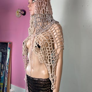 May include: A handmade crochet hooded shawl in shades of beige, gray, and pink. The open-weave design features a hood and a draped neckline. The mannequin also wears a black and pink crochet skirt.