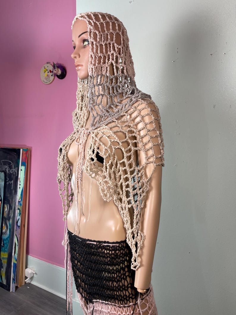 May include: A handmade crochet hooded shawl in shades of beige, gray, and pink. The open-weave design features a hood and a draped neckline. The mannequin also wears a black and pink crochet skirt.