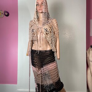 May include: A handmade crochet outfit on a mannequin. The outfit includes a hooded shawl and a skirt. The shawl is beige and the skirt is a patchwork of black, pink, and beige crochet. The skirt has fringe details.