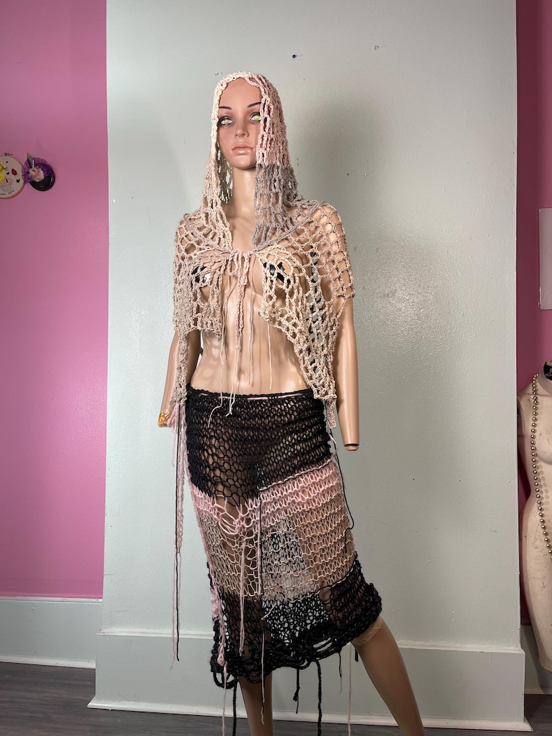 May include: A handmade crochet outfit on a mannequin. The outfit includes a hooded shawl and a skirt. The shawl is beige and the skirt is a patchwork of black, pink, and beige crochet. The skirt has fringe details.