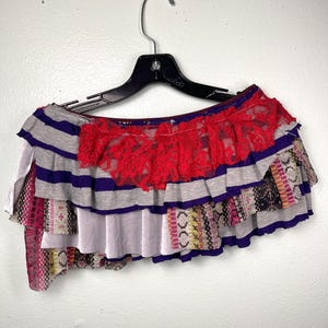 Puede incluir: Una falda de volantes superpuestos con rayas horizontales grises y moradas, un borde de encaje rojo vibrante en la parte superior y una sección inferior de tela estampada multicolor. La prenda se muestra en una percha de plástico negra.
