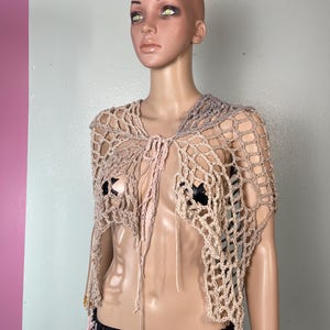 May include: A beige and gray crocheted shawl with a net-like design. The open-weave garment has a tie closure and is displayed on a mannequin. The shawl is a unique, handmade fashion accessory.