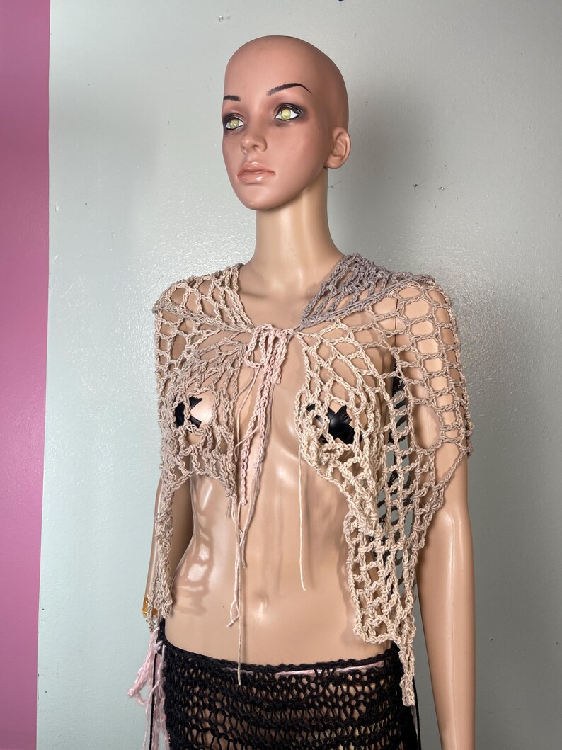 May include: A beige and gray crocheted shawl with a net-like design. The open-weave garment has a tie closure and is displayed on a mannequin. The shawl is a unique, handmade fashion accessory.