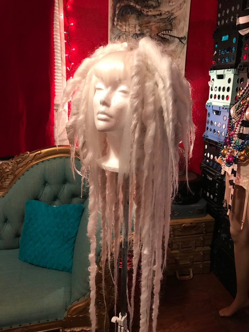 Puede incluir: Una peluca blanca con rastas largas y gruesas. La peluca est&aacute; en una cabeza de poliestireno y est&aacute; sentada en un soporte.