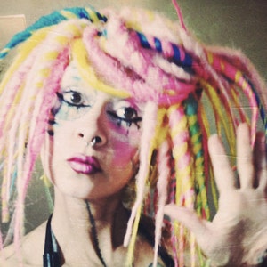 Könnte beinhalten: Eine Person mit pinken, gelben und blauen Dreadlocks, die farbenfrohes Make-up und einen Nasenring trägt. Die Person trägt ein schwarzes Halstuch und hat die Hand erhoben.