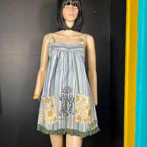 May include: A sleeveless dress featuring vertical blue, green, and white stripes with a gathered neckline. The dress includes two front pockets with a yellow and white patterned print, a central graphic of two figures and a sun, and a dark green ruffled hem.
