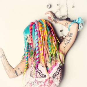 Puede incluir: Una mujer con rastas de color azul brillante y multicolor, lleva un vestido floral rosa y blanco con ribetes de encaje negro. Tiene varios tatuajes en los brazos, incluyendo una ballena con n&uacute;meros romanos.