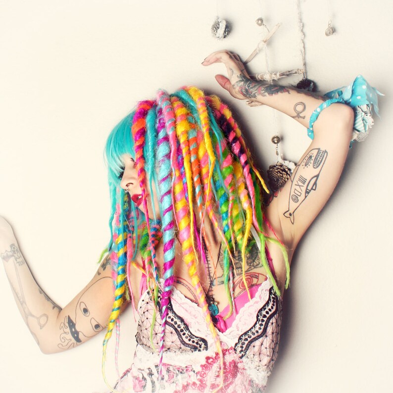 Puede incluir: Una mujer con rastas de color azul brillante y multicolor, lleva un vestido floral rosa y blanco con ribetes de encaje negro. Tiene varios tatuajes en los brazos, incluyendo una ballena con n&uacute;meros romanos.