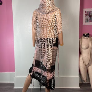 May include: A handmade, open-weave crochet garment in shades of pink, black, and beige, displayed on a mannequin. The garment appears to be a hooded shawl and skirt, with a loose, fringed hemline. The design features a large, open net-like pattern.
