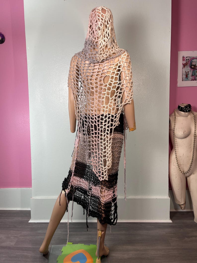 May include: A handmade, open-weave crochet garment in shades of pink, black, and beige, displayed on a mannequin. The garment appears to be a hooded shawl and skirt, with a loose, fringed hemline. The design features a large, open net-like pattern.