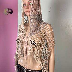 May include: A beige and gray crocheted hooded shawl with a net-like design. The shawl has a draped, open front and long, dangling fringe. The mannequin is also wearing a black skirt.