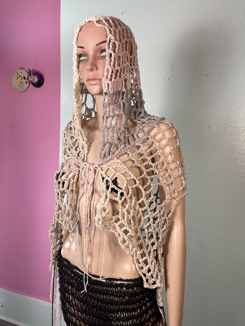 May include: A beige and gray crocheted hooded shawl with a net-like design. The shawl has a draped, open front and long, dangling fringe. The mannequin is also wearing a black skirt.