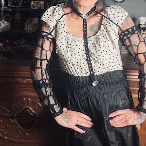 May include: A woman wearing a white and black polka dot dress with black crocheted sleeves and a black crocheted choker necklace.