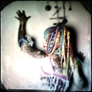 Puede incluir: Una mujer con rastas de colores y tatuajes lleva una blusa blanca con un estampado colorido. Su brazo est&aacute; levantado y su mano est&aacute; abierta.