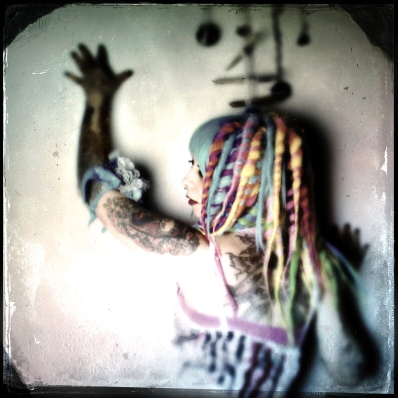 Puede incluir: Una mujer con rastas de colores y tatuajes lleva una blusa blanca con un estampado colorido. Su brazo est&aacute; levantado y su mano est&aacute; abierta.