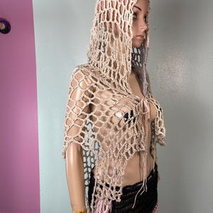 May include: A beige crocheted hooded shawl with a net-like design. The shawl is draped over a mannequin, showcasing its open weave and flowing shape. The garment has a tie closure at the front and is paired with a black and pink skirt.