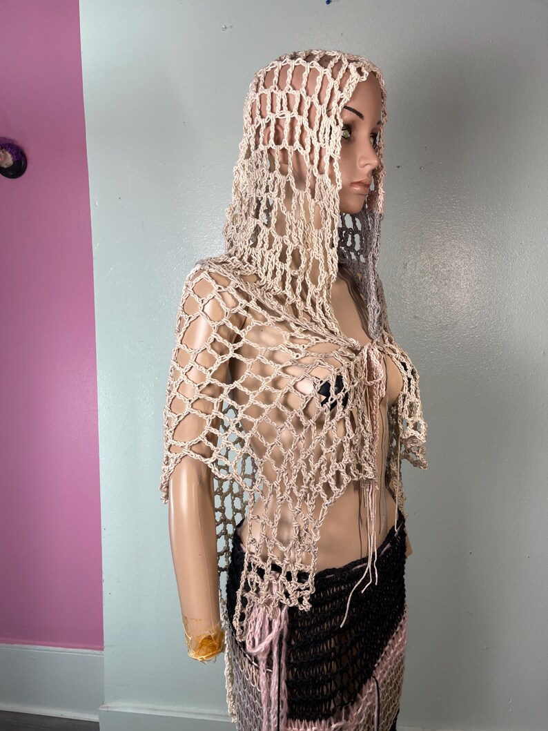 May include: A beige crocheted hooded shawl with a net-like design. The shawl is draped over a mannequin, showcasing its open weave and flowing shape. The garment has a tie closure at the front and is paired with a black and pink skirt.